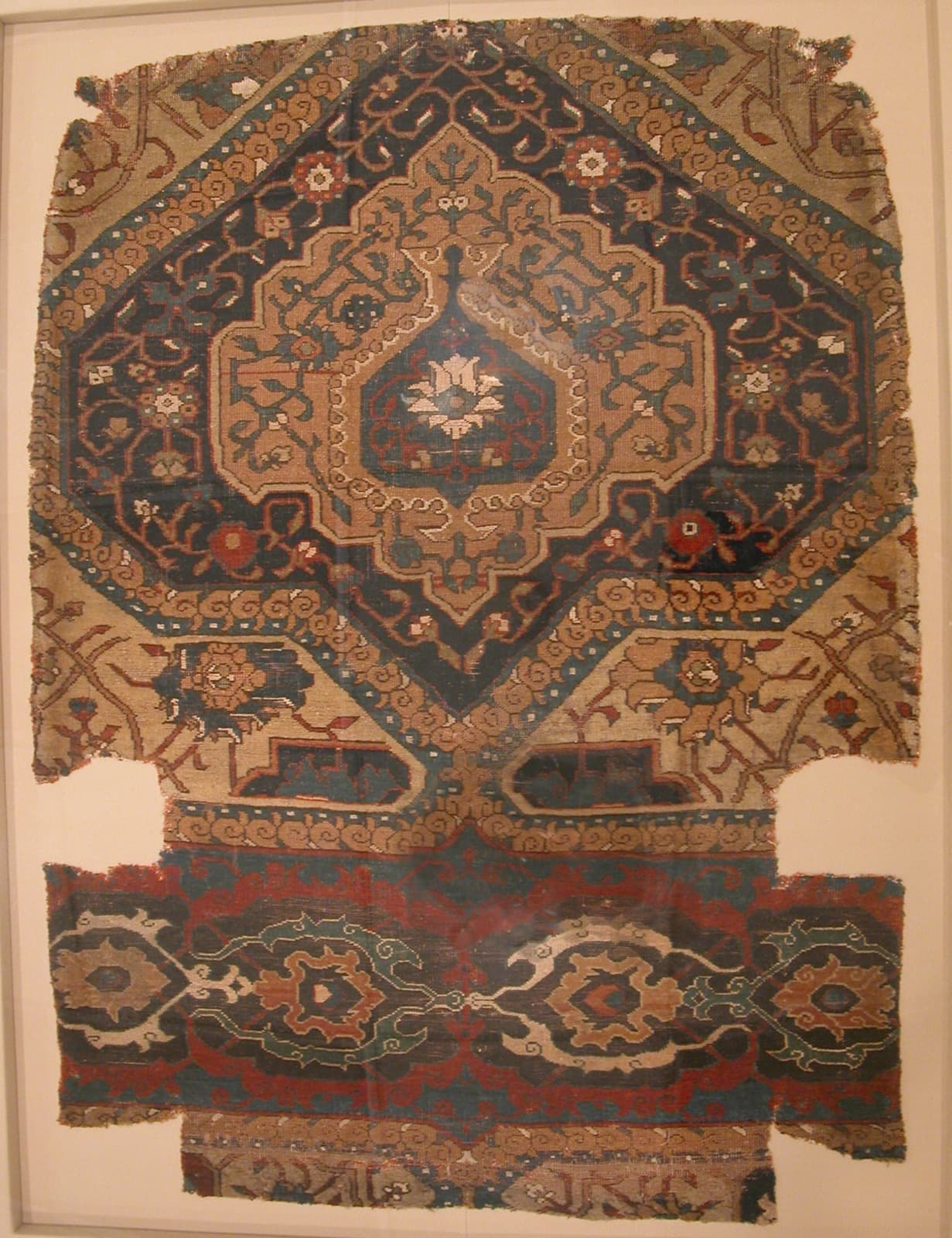 A worn carpet fragment mounted for display: a stepped central medallion in indigo and cream above a red and blue border band, the edges frayed.