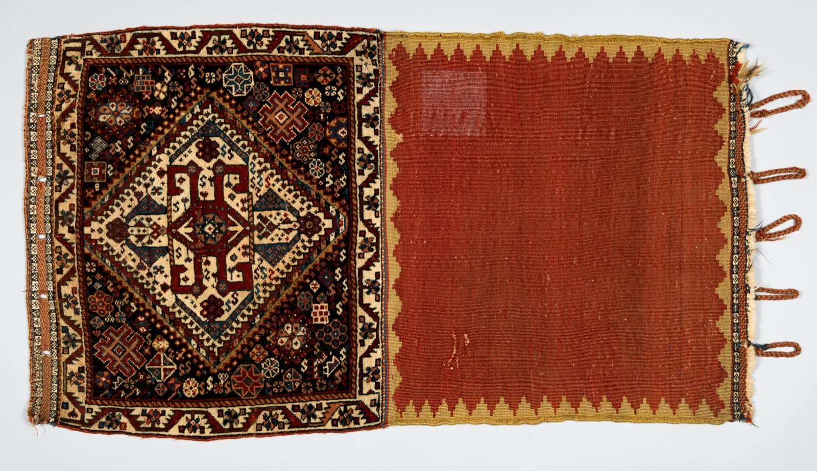 Half a saddle bag laid flat: a knotted pile face with a stepped cream and red medallion on the left, and a plain red woven back with a tasselled edge on the right.